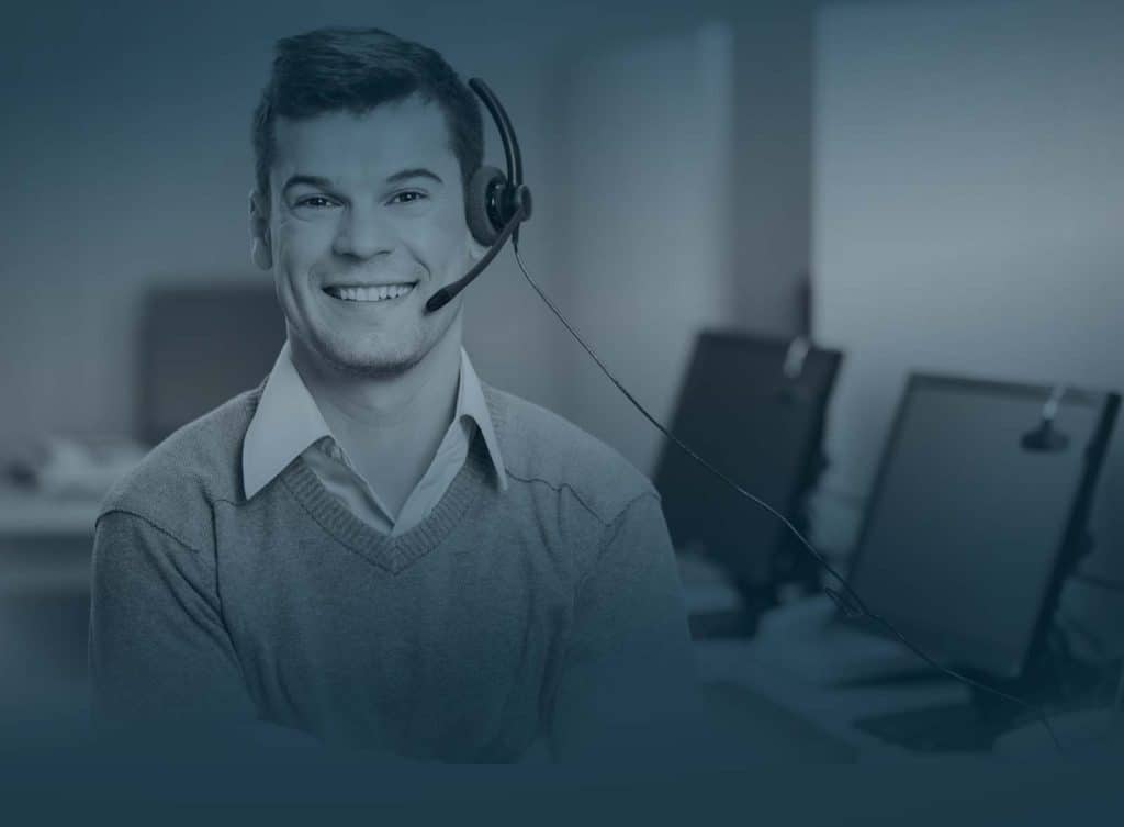 Ein lächelnder Mann mit Headset in einem Büro, mit Computerbildschirmen im Hintergrund.
