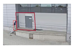 Eine Person verlässt ein Gebäude durch eine Tür auf einer erhöhten Plattform, umgeben von roten Umrisslinien.