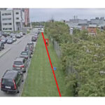 Bild einer städtischen Szene mit geparkten Autos entlang einer Straße. Eine rote Linie verläuft diagonal über eine grasbewachsene Fläche. Im Hintergrund sind Gebäude zu sehen.