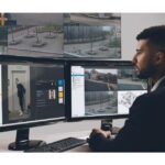 Ein Mann sitzt vor mehreren Computerbildschirmen, die Überwachungsbilder und Sicherheitssoftware zeigen.