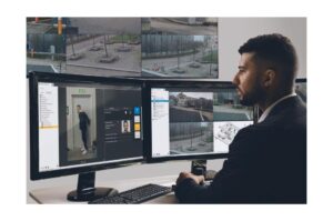 Ein Mann sitzt vor mehreren Computerbildschirmen, die Überwachungsbilder und Sicherheitssoftware zeigen.