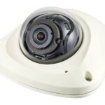 Überwachungskamera in einer weißen, dreieckigen Halterung mit transparentem Schutzgehäuse.