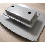 Ein grauer Metallkasten mit zwei Löchern auf einer rechteckigen Basis, auf einem Holztisch platziert.