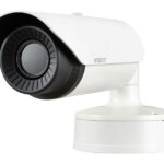 Weiße Überwachungskamera mit schwarzem Objektiv auf einem Sockel.