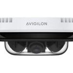 Eine weiße Überwachungskamera von Avigilon in einer Seitenansicht, erkennbar am transparenten Gehäuse, das die Kameraoptik zeigt.
