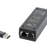 Ein schwarzer Netzwerkadapter mit zwei Ethernet-Anschlüssen und einem USB-Dongle daneben. Auf der Oberseite des Geräts befinden sich technische Informationen.