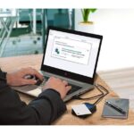 Ein Mann arbeitet an einem Laptop an einem Schreibtisch mit einem USB-Stick und einem Notizbuch daneben.