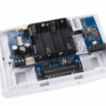 Elektronisches Gerät mit offenen Gehäuse auf einem weißen Kunststoffrahmen montiert, zeigt eine blaue Leiterplatte, Batteriefach und elektronische Komponenten.