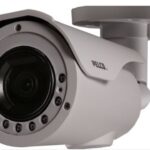 Nahaufnahme einer weißen Überwachungskamera mit zylindrischem Gehäuse und sichtbarem Objektiv, an der Wand montiert.