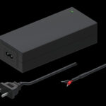 Rechteckiges schwarzes Netzteil mit einem grünen Indikatorlicht auf der Oberseite, einem Stromkabelanschluss und zwei separaten Kabeln, eines mit einem Stecker und das andere mit offenen Kabelenden.