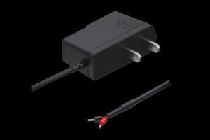 Schwarzes Netzteil mit zwei Flachsteckern und einem Kabel, das mit einem roten und einem schwarzen Stecker endet, auf schwarzem Hintergrund dargestellt.