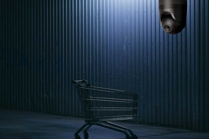 Ein Einkaufswagen steht vor einer dunklen, metallischen Wand, und eine Überwachungskamera ist im oberen Bildrand sichtbar.