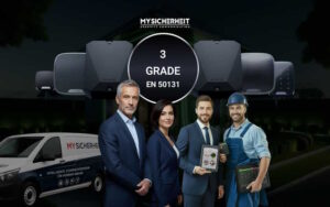 Eine Gruppe von vier Personen, darunter ein Geschäftsmann, eine Geschäftsfrau, ein Techniker mit einem Tablet und ein Handwerker, steht vor einem Hintergrund mit technischen Geräten und einem Lieferwagen mit der Aufschrift 'My Sicherheit'. Im Zentrum des Bildes sind die Worte '3 Grade EN 50131' zu sehen.