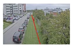 Bild einer städtischen Szene mit geparkten Autos entlang einer Straße. Eine rote Linie verläuft diagonal über eine grasbewachsene Fläche. Im Hintergrund sind Gebäude zu sehen.