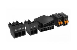 Schwarze elektrische Steckverbinder mit nummerierten Anschlüssen und Beschriftungen: "NO, COM, NC" und "+, -", daneben Steckplätze für Kabel.