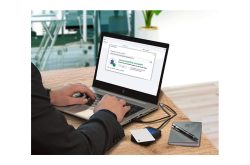 Ein Mann arbeitet an einem Laptop an einem Schreibtisch mit einem USB-Stick und einem Notizbuch daneben.