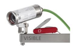 Eine silberne Kamera mit einem grünen Kabel, montiert auf einer Metallhalterung mit der Aufschrift "VISIBLE", daneben ein rotes Taschenmesser mit ausgeklappter Klinge.